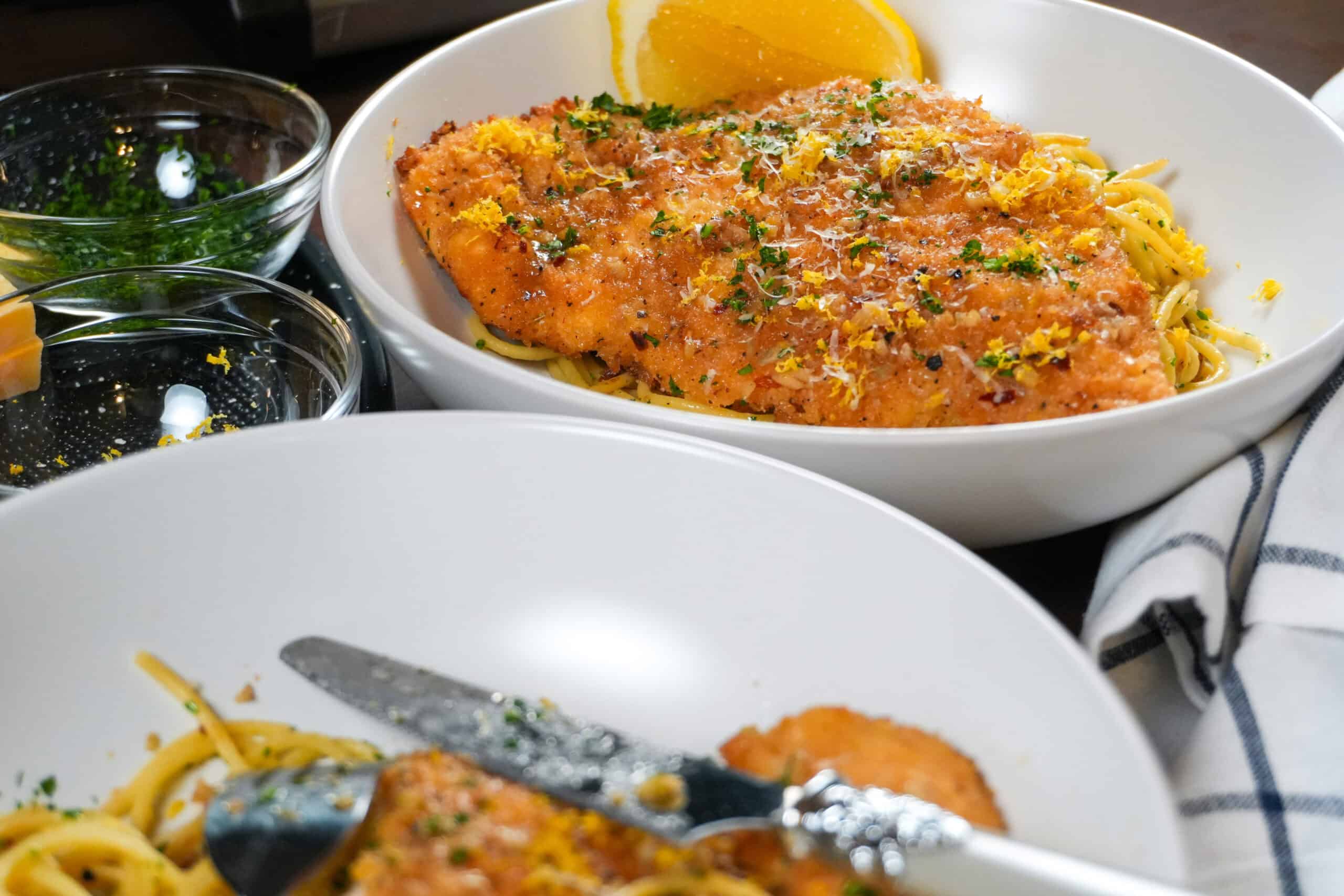 Elegant chicken cutlet garnished with herbs, served with pasta and lemon in a refined dining setting.